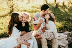 family of five sits on a log mom kisses toddler an dad kisses older girl
