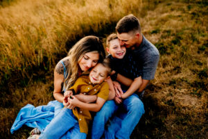 Portland Family Photographer, mother, dad and two sons each parent has a child on their lap as they sit on the floor some have their eyes closed and some are smiling