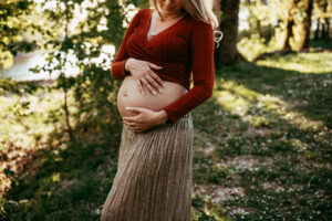 Maternity Photographer, a pregnant mother holds her belly beneath the shade of trees on a sunny