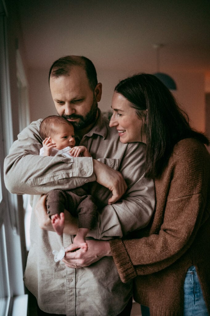 Newborn Photographer, mom and dad hold baby by the window in their home in Portland